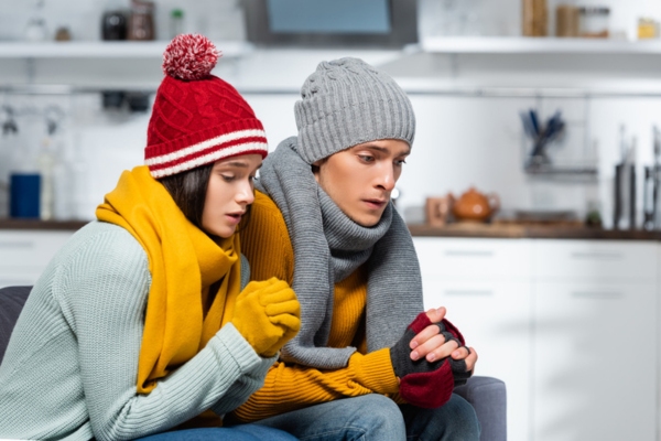 Young couple in warm hats, scarfs and gloves sitting on a couch depicting furnace failure